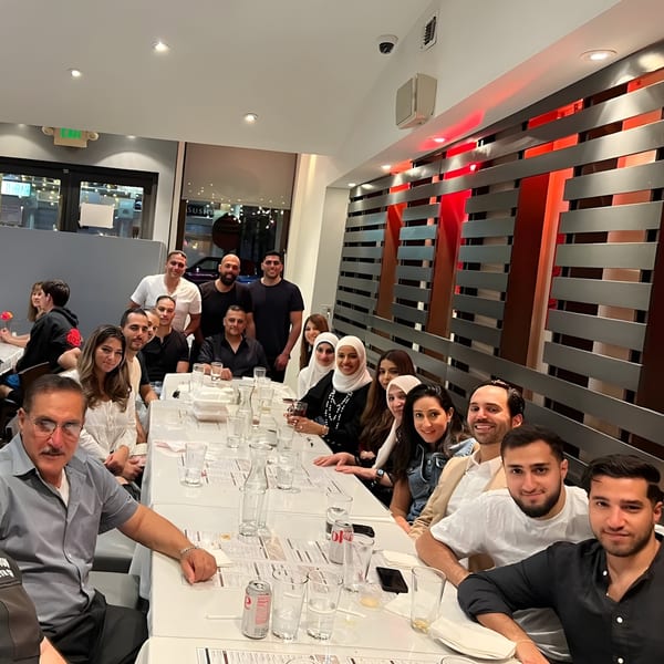 A large group of people, men and women, seated at long tables in Farsi Cafe, smiling.