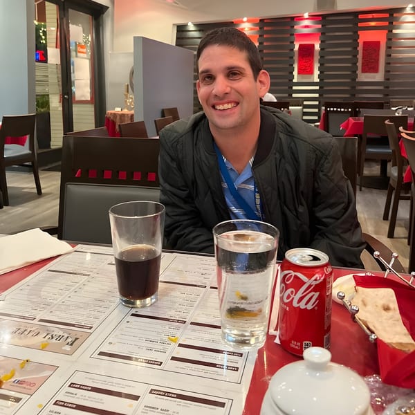 Smiling man seated at a Farsi Cafe table with drinks and a menu, red decor in background.